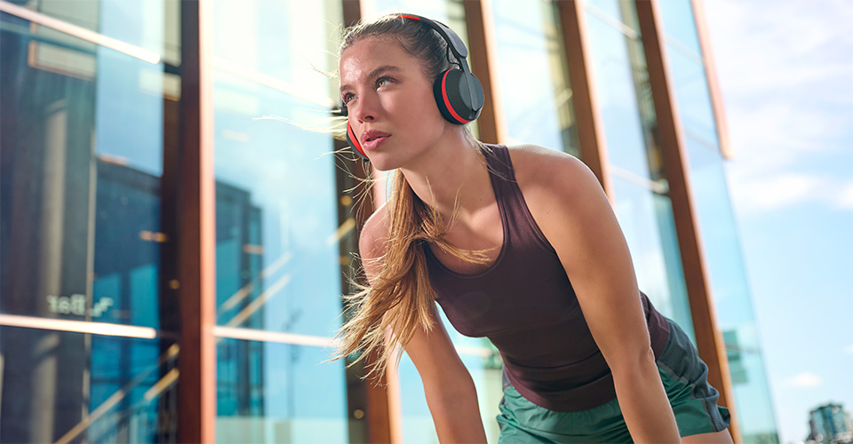 Une femme portant un casque Philips A6219 fait une pause lors d'une s&eacute;ance d'entra&icirc;nement en plein air, debout devant un b&acirc;timent en verre moderne