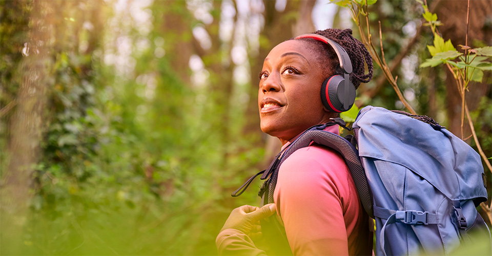 Une femme fait de la randonn&eacute;e dans une for&ecirc;t avec un sac &agrave; dos bleu, portant un casque Philips A6219 et levant les yeux avec un sourire