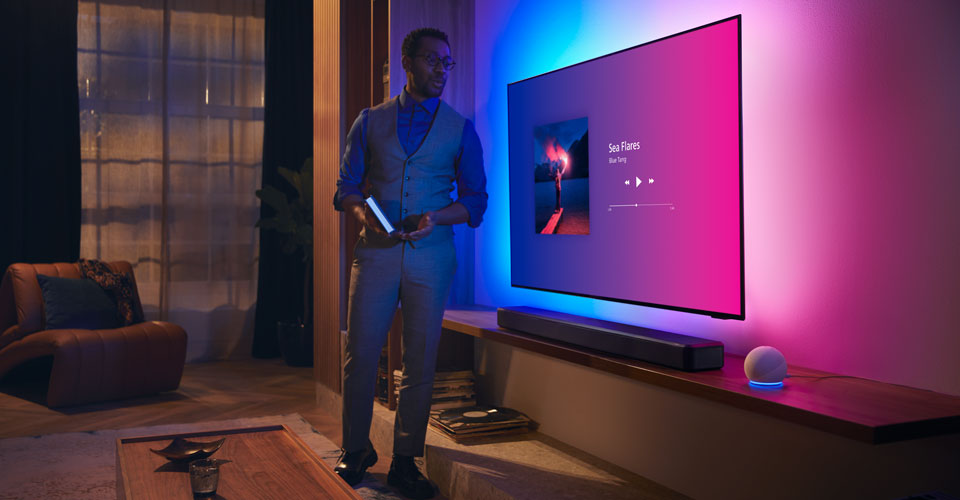 Personne dans un salon moderne pr&egrave;s d'un t&eacute;l&eacute;viseur Philips Ambilight avec interface musicale et barre de son Fidelio