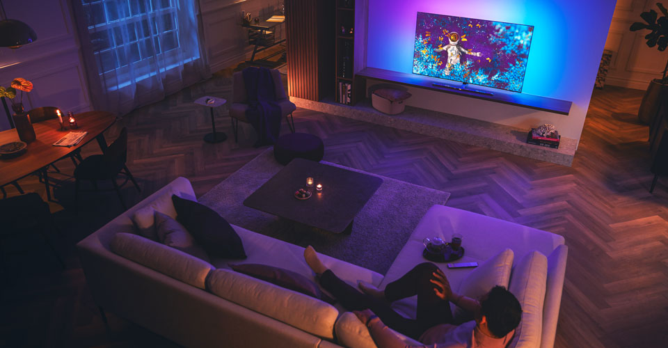 Un homme se d&eacute;tend sur un canap&eacute; dans un salon faiblement &eacute;clair&eacute;, regardant un t&eacute;l&eacute;viseur Philips Ambilight