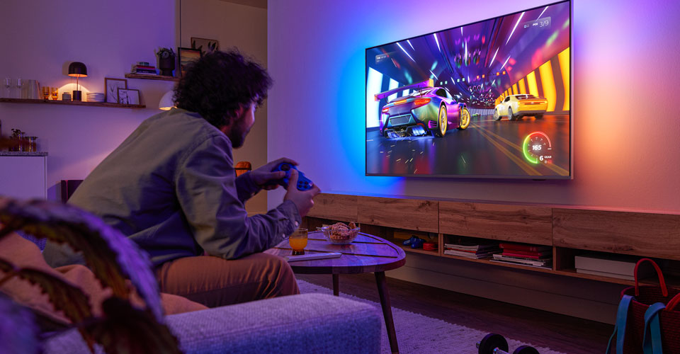 Une personne est assise sur un canap&eacute; et joue &agrave; un jeu vid&eacute;o de course sur un t&eacute;l&eacute;viseur Philips Ambilight